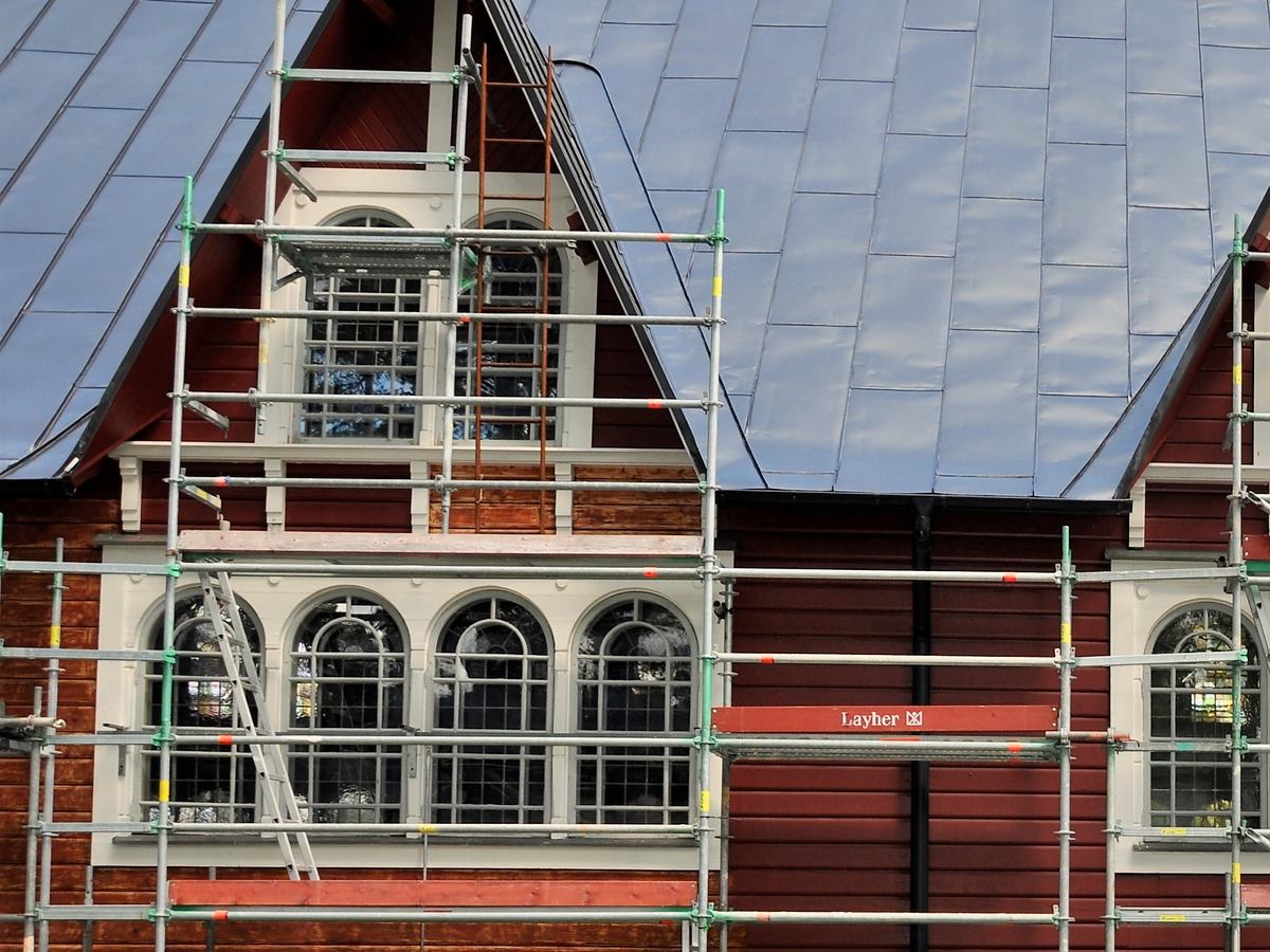Gaveln på ett rött trähus med spröjsade fönster och byggnadsställning under ett fasadjobb