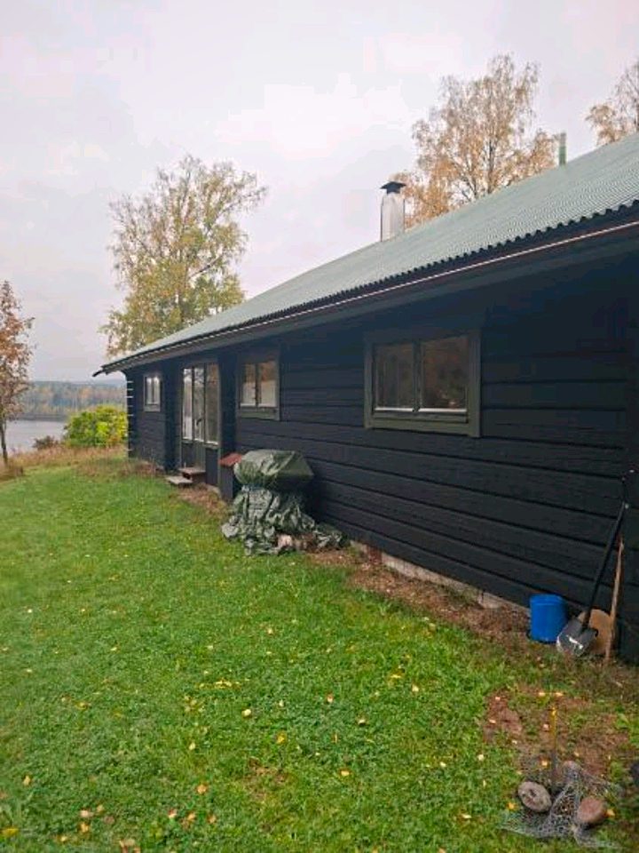 Långsidan på ett svart timmerhus med gröngrå fönster och en sjö i bakgrunden